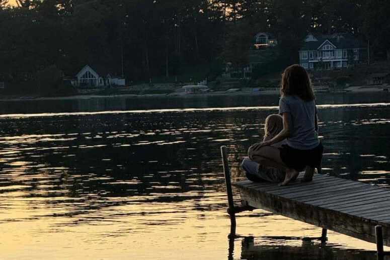 A Woman And Child Sit On Dock Looking At Glistening Lake At Sunset.