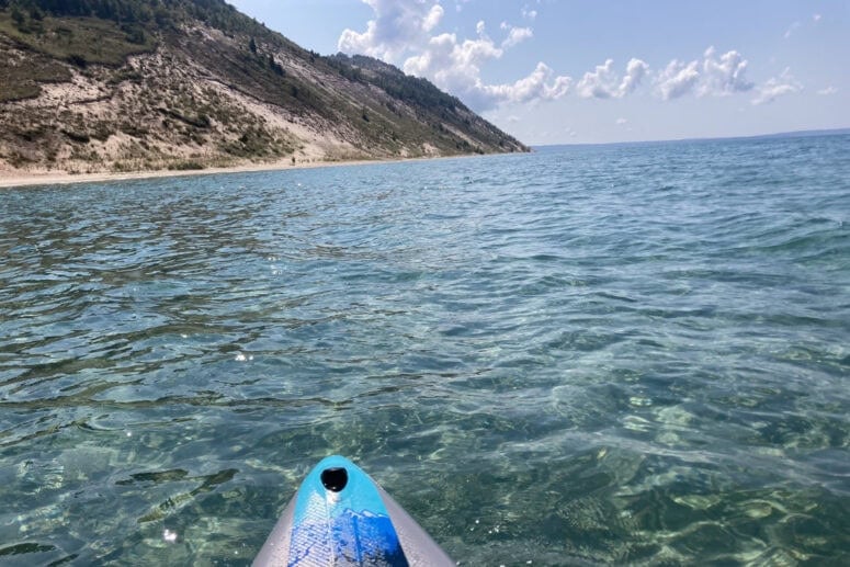 A Paddleboard Bobs In The Water Looking At A Sandy Dune.